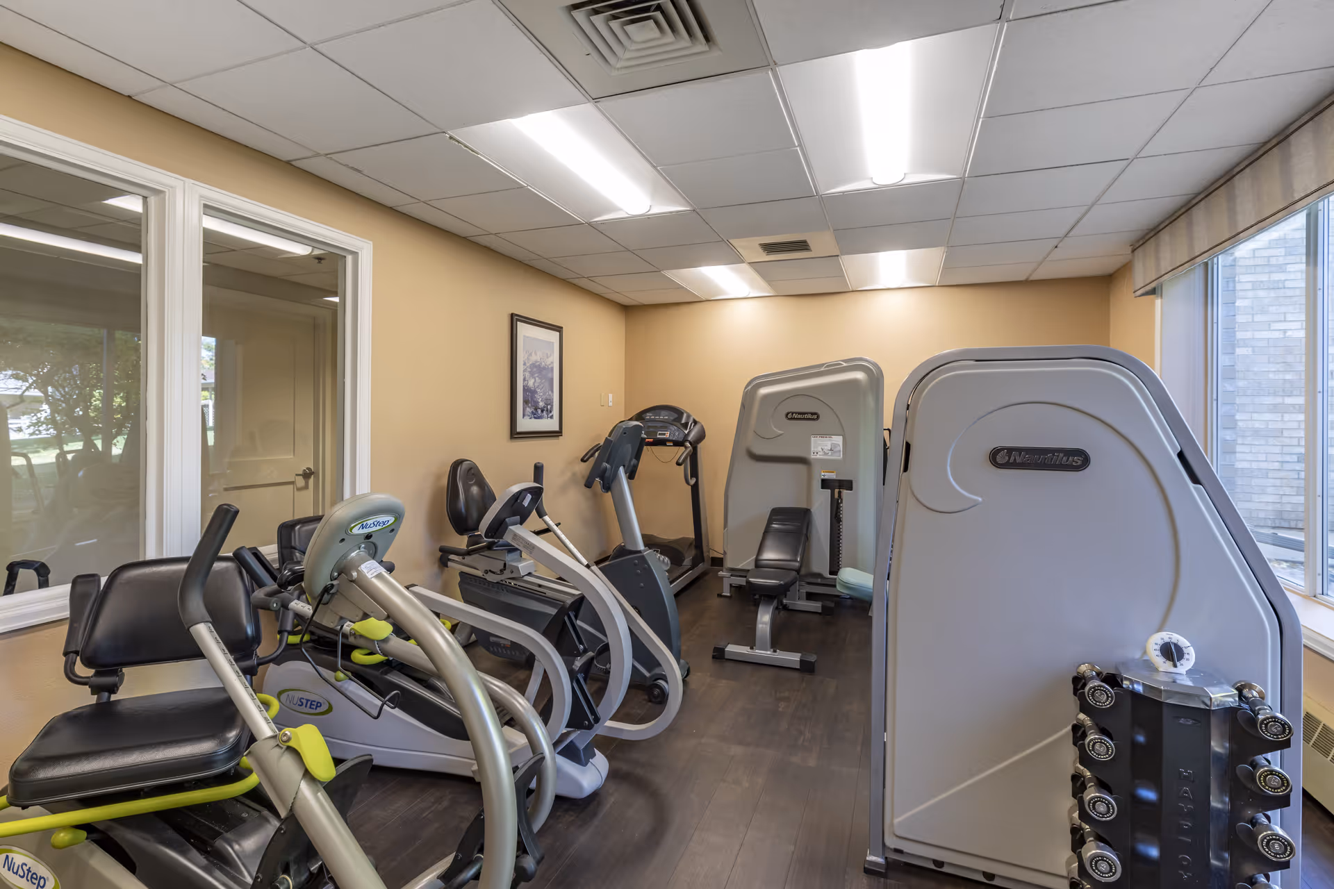 A small fitness room with exercise equipment including a recumbent bike, a treadmill, and weight machines. The room has beige walls, a dark floor, fluorescent ceiling lights, a framed picture on the wall, and a large window letting in natural light.