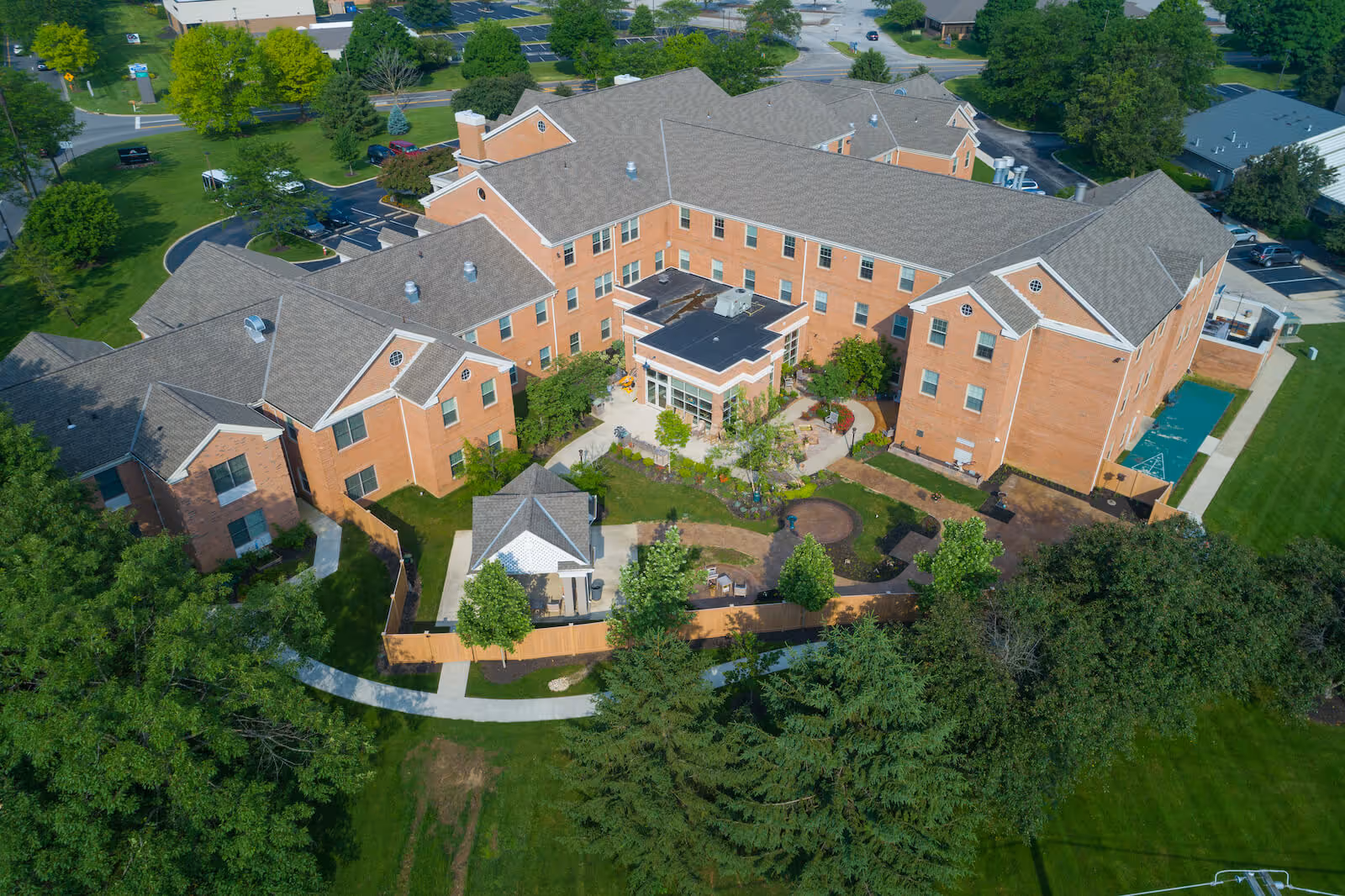 Aerial view of a large senior living facility named Harmony Trace, featuring a multi-wing brick building with a central courtyard, surrounded by green lawns, trees, and paved walkways.