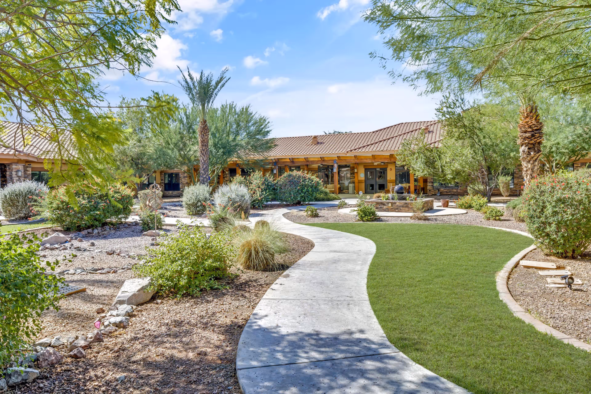 A curved concrete path winds through desert landscaping toward the front entrance of a single-story senior living building.