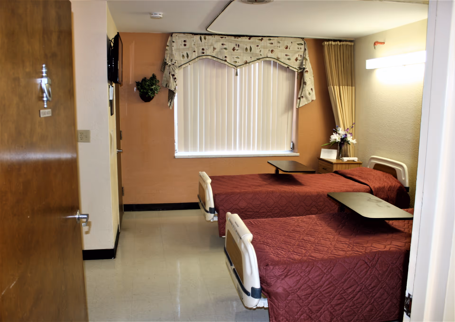 A nursing facility bedroom with two hospital beds covered in maroon quilts, each with an overbed table. There is a window with vertical blinds and a decorative valance, a small nightstand with a flower arrangement, and a wall-mounted light fixture.