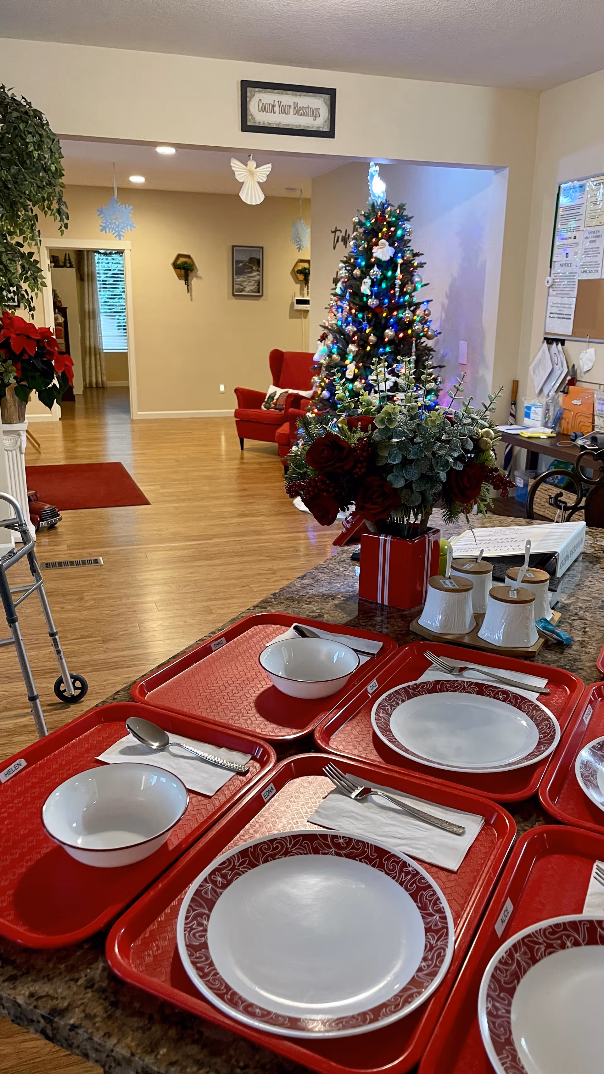 Interior view of a senior living facility dining area with red trays set on a counter, each tray holding a plate, bowl, napkin, and utensils. In the background, there is a decorated Christmas tree with colorful lights, a red armchair, and holiday decorations hanging from the ceiling. A walker is visible on the left side, and a bulletin board with papers is on the right wall.