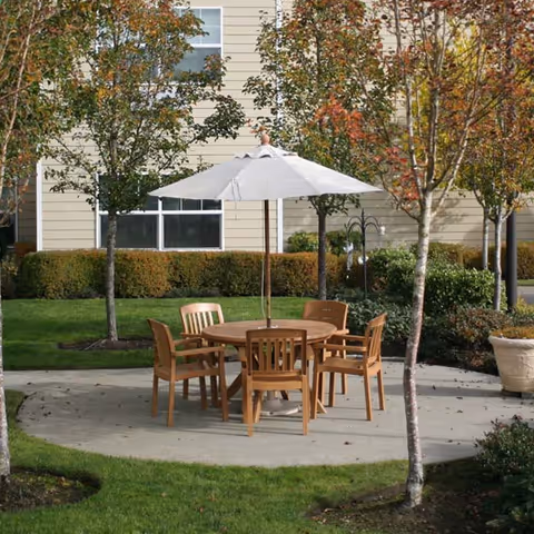 Outdoor patio area with a round wooden table and six matching wooden chairs arranged around it. A large white umbrella is centered over the table. The patio is surrounded by green grass, small trees with autumn-colored leaves, and shrubs. A beige building with windows is visible in the background.