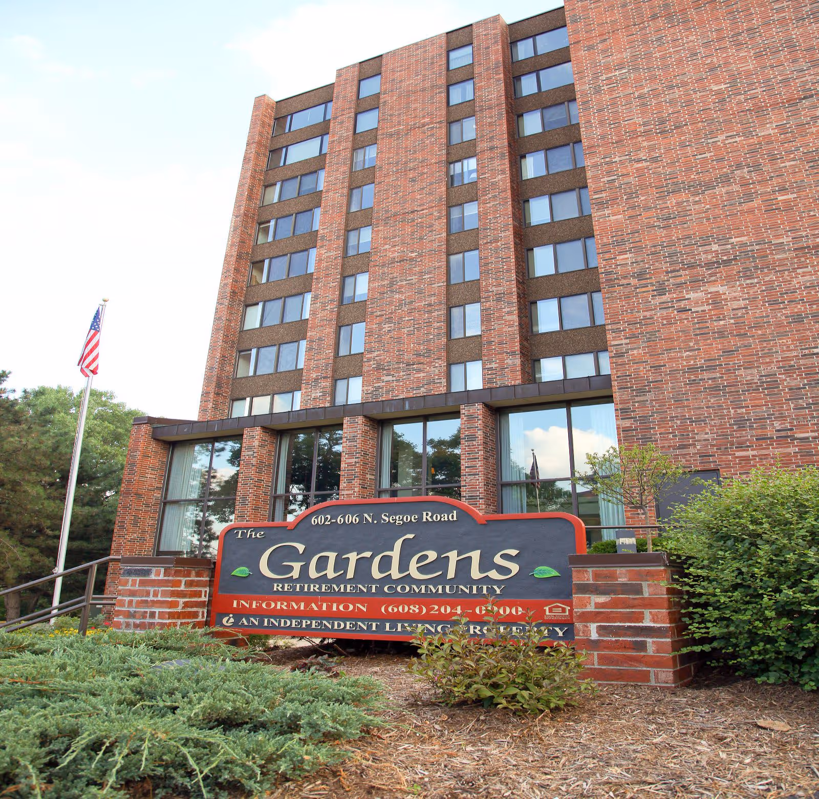 Exterior view of The Gardens Retirement Community building, a multi-story brick structure with many windows. In front of the building is a large sign displaying the community name, address, and contact information, surrounded by greenery and an American flag on a flagpole to the left.