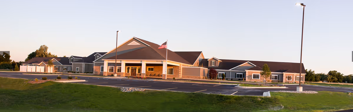 Exterior view of The Meadows of Owosso senior living facility building with a large parking lot, green lawn, and an American flag on a flagpole in front of the entrance during daylight.