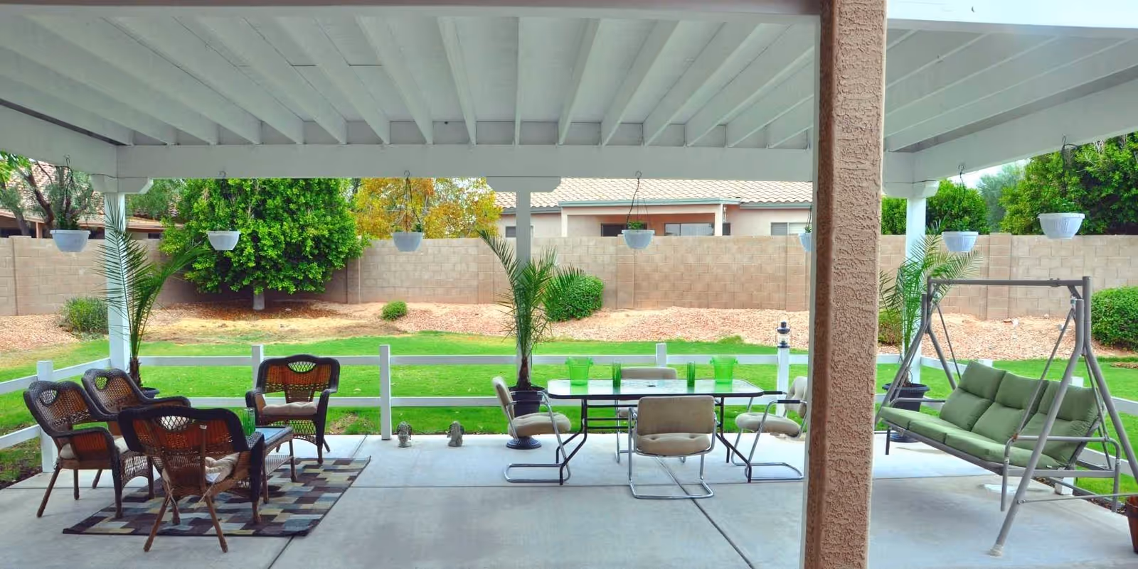 Covered backyard patio with multiple seating areas, a dining table, green cushioned swing, potted plants, and a lawn with a block wall beyond.