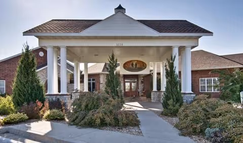 Front entrance of a brick senior living building with a covered portico supported by white columns and surrounding landscaping.