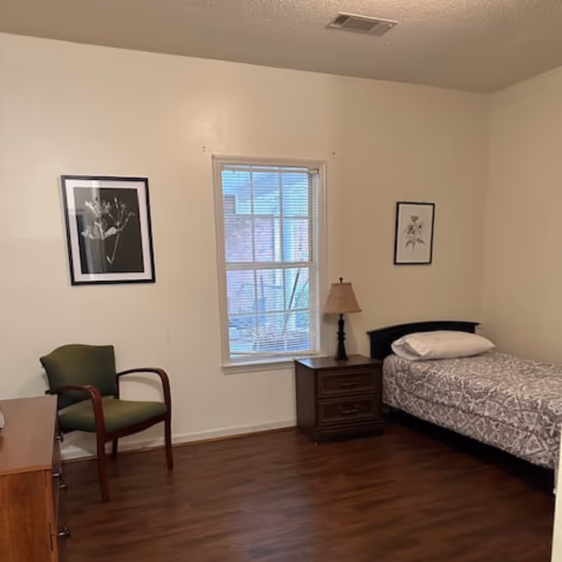 A simple bedroom with a single bed, nightstand and lamp, a green chair, framed wall art, and a window.