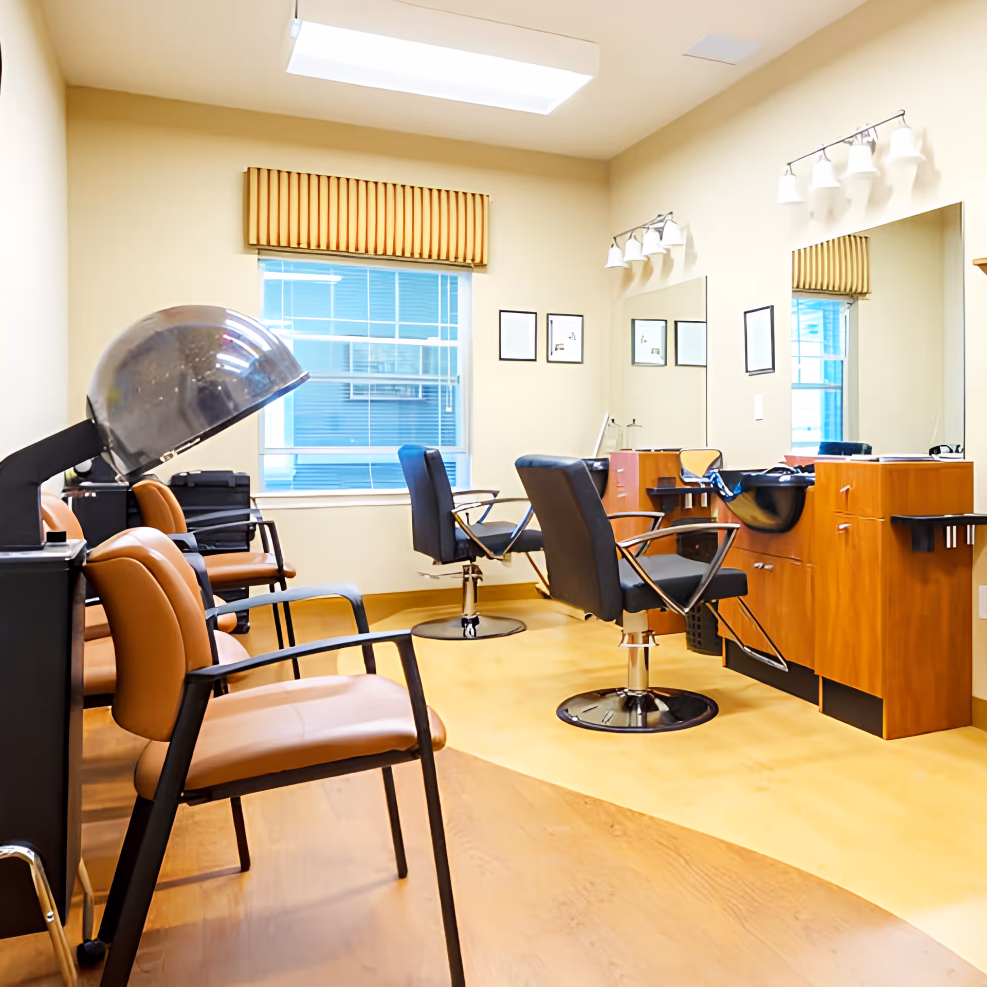 Interior of a hair salon area with salon chairs, a hair dryer, large mirrors, and wooden cabinetry under bright lighting. There is a window with a striped valance and framed pictures on the wall.