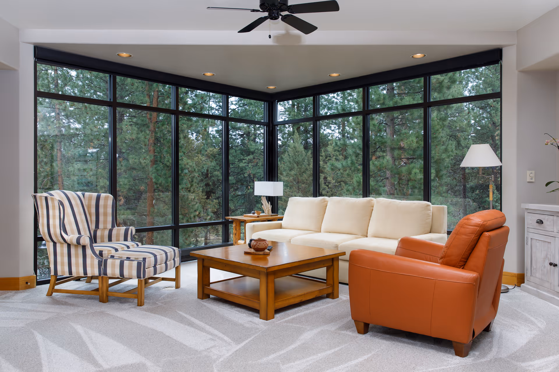 Bright living room with floor-to-ceiling windows, a cream sofa, orange armchair, striped wingback chair, and a wooden coffee table.