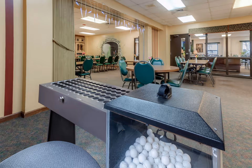 Interior view of a senior living facility common area with multiple round tables and green chairs arranged for dining or social activities. In the foreground, there is a bingo ball machine filled with white bingo balls. The room has beige walls, carpeted floors, and fluorescent ceiling lights. A mural of a stone archway is visible on the far wall.