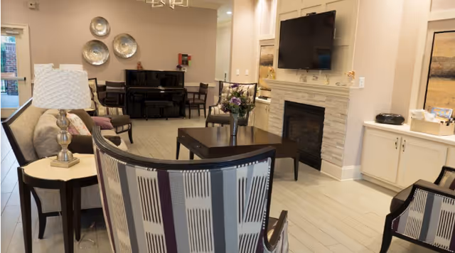 A cozy living room area in an assisted living facility featuring a fireplace with a mounted flat-screen TV above it, a dark wooden coffee table with a vase of flowers, several upholstered chairs with striped patterns, a side table with a lamp, and a black piano against the far wall with decorative silver plates hanging above it.