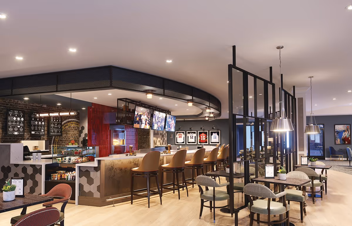 A modern dining and lounge area with a bar counter featuring high chairs, multiple TV screens mounted above the bar, and a coffee and snack station with a menu on the wall. The space has a mix of seating options including tables with chairs and pendant lighting, with sports jerseys framed on the wall in the background.