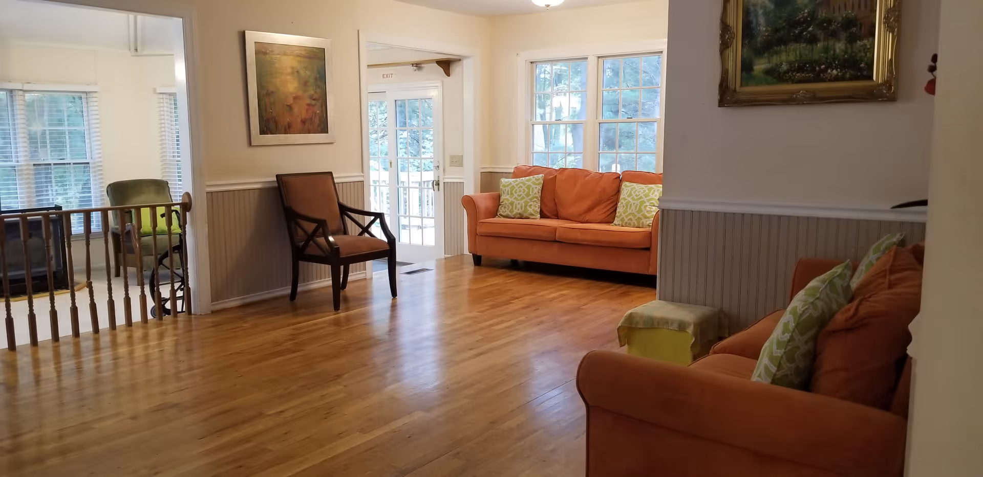 A bright living room with wooden floors featuring two orange sofas with green patterned pillows, a small green ottoman, a wooden chair, and large windows letting in natural light. There is a painting on the wall and a door leading outside.