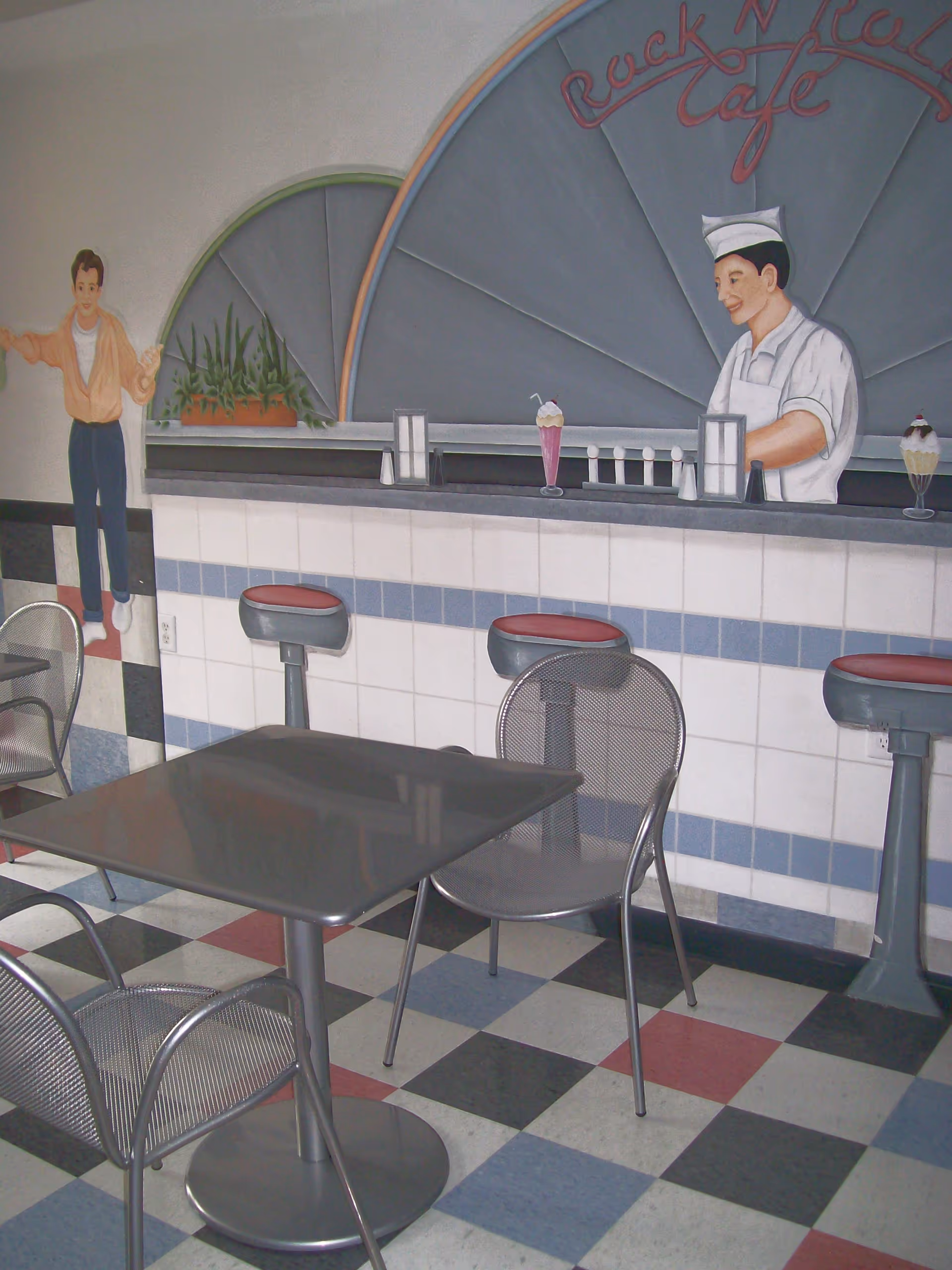 Interior of a retro-style cafe with a checkered floor in black, white, red, and blue tiles. There are metal tables and chairs in the foreground. The back wall features a mural of a soda jerk behind a counter with stools, milkshakes, and a young boy. The mural includes the text 'Rock N Roll Cafe'.