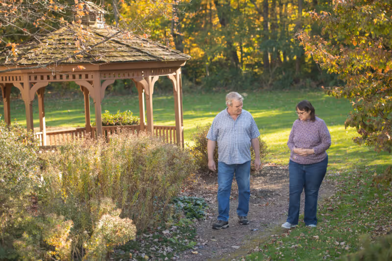 An elderly man and woman walking and talking on a garden path next to a wooden gazebo surrounded by greenery and trees with autumn foliage.