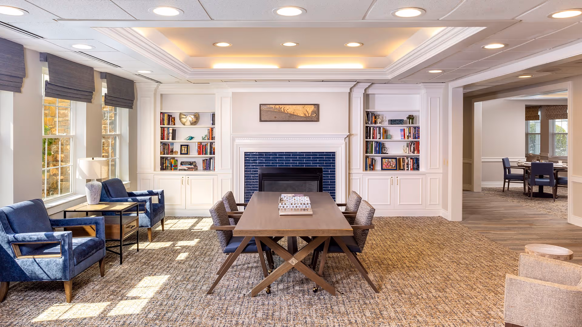 A bright and spacious living room area with a central wooden table surrounded by six chairs. The room features a fireplace with blue tiles and built-in white bookshelves on either side filled with books and decorative items. To the left, there are three large windows with gray roman shades and two blue upholstered armchairs with a side table and lamp between them. The floor is carpeted and there is an adjacent room visible through an open doorway with additional seating and dining tables.