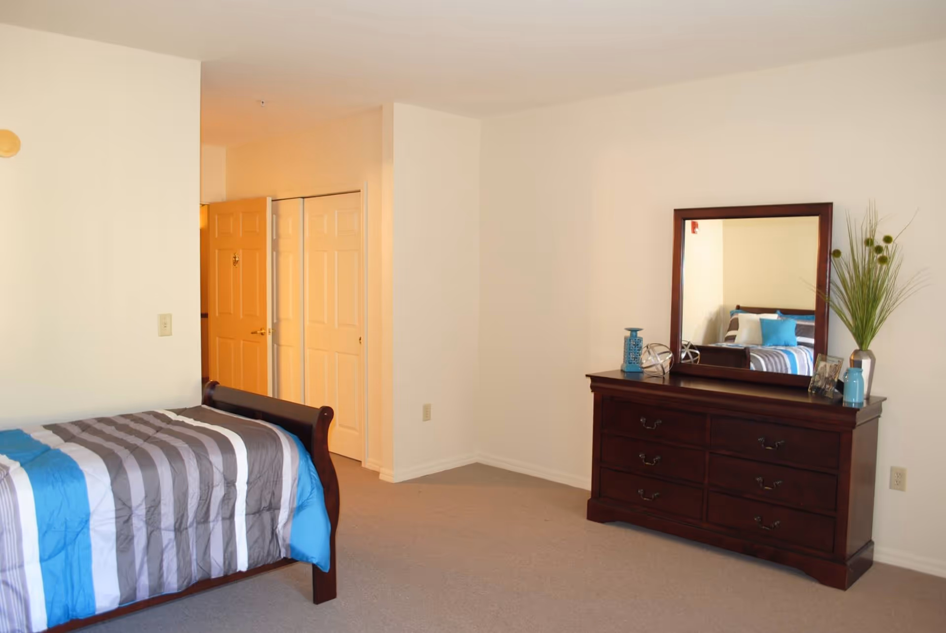 A bedroom with a wooden bed frame and a striped blue, gray, and white comforter. There is a wooden dresser with six drawers and a large mirror on top. The dresser is decorated with a vase of tall green plants, a blue lantern, a small blue jar, and a photo frame. The room has beige carpet and white walls, with a closet and an open door visible in the background.