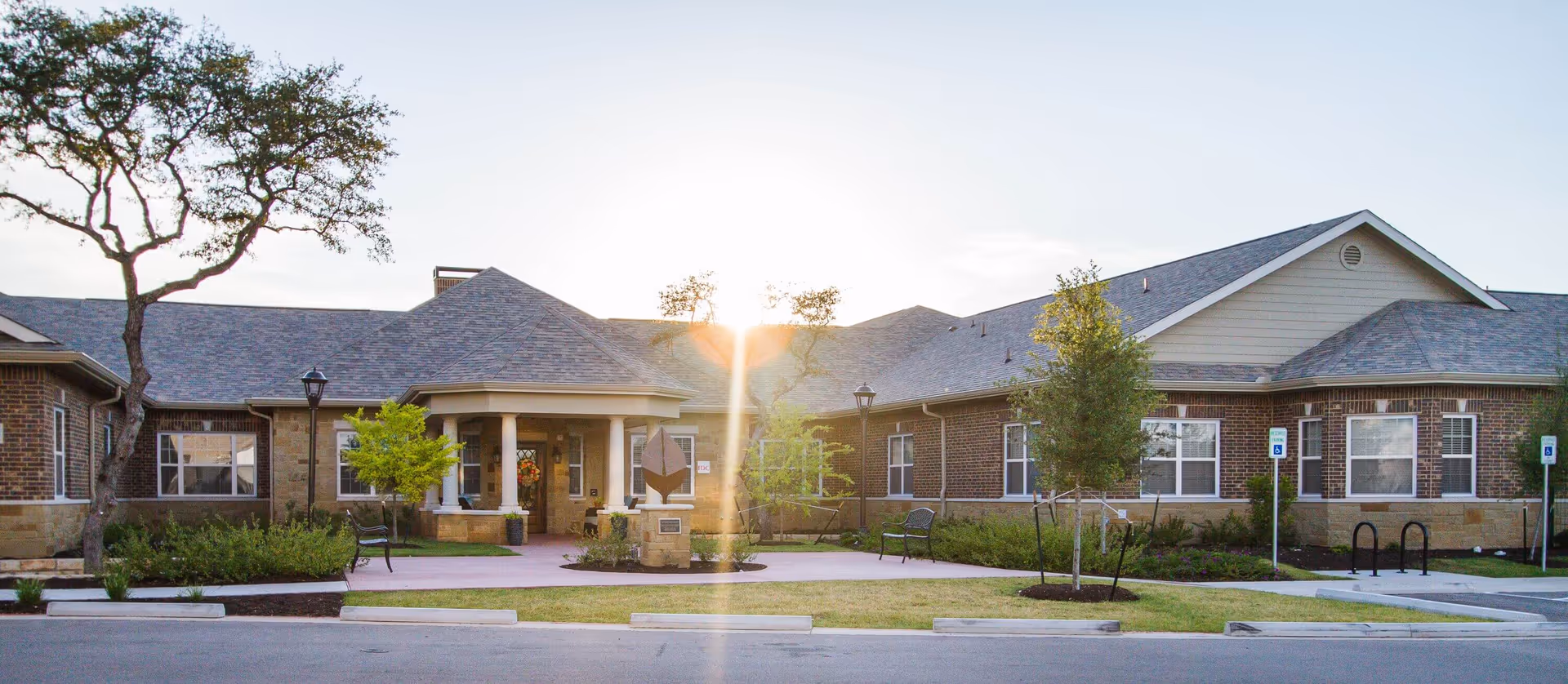 Front exterior view of a single-story brick building with a covered entrance, surrounded by small trees, benches, and a parking area with handicapped parking signs. The sun is setting behind the building.
