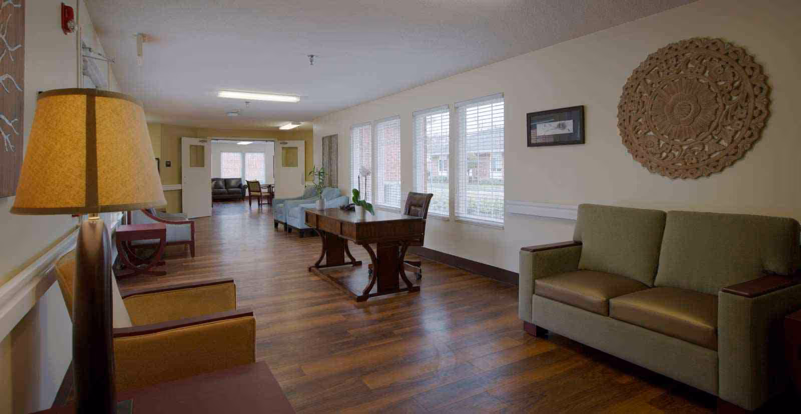 A well-lit interior common area with wooden flooring, featuring multiple seating options including armchairs and sofas. A wooden table with a chair is positioned near large windows with blinds. Decorative wall art and a lamp are also visible.