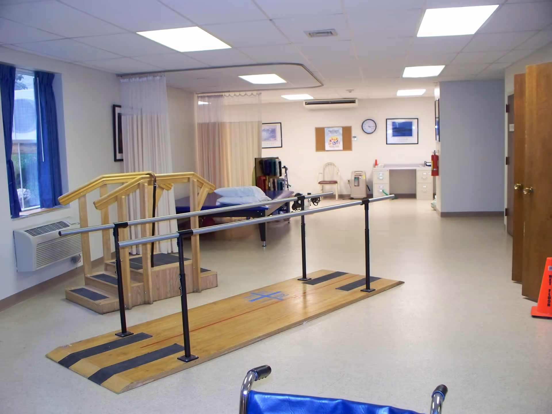 A rehabilitation room with parallel bars for walking exercises, a small set of wooden stairs with handrails, a treatment table with pillows, and various medical equipment. The room has white walls, a clock, framed pictures, and a desk in the background.