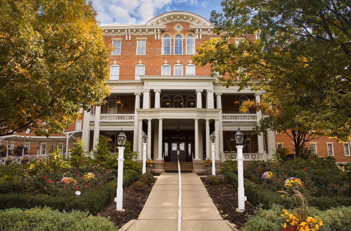 Front exterior view of a large, multi-story brick building with white columns and a covered entrance. The building is surrounded by well-maintained gardens with colorful flowers and trees with autumn foliage. A paved walkway leads up to the entrance, flanked by white lamp posts.