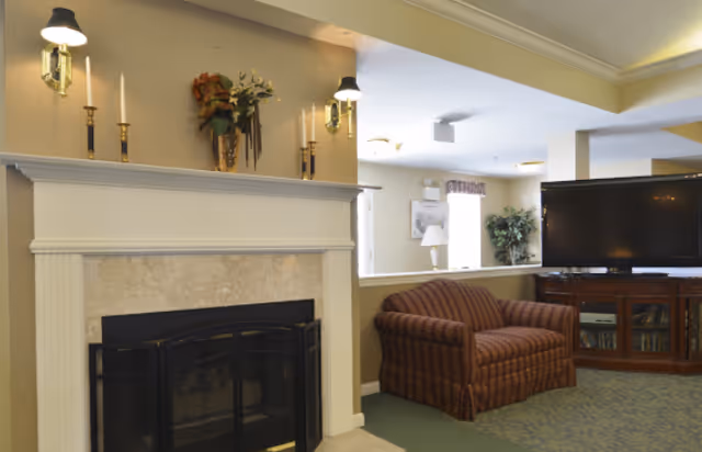 A cozy common room featuring a fireplace, wall sconces, a striped armchair, and a large flat-screen TV.