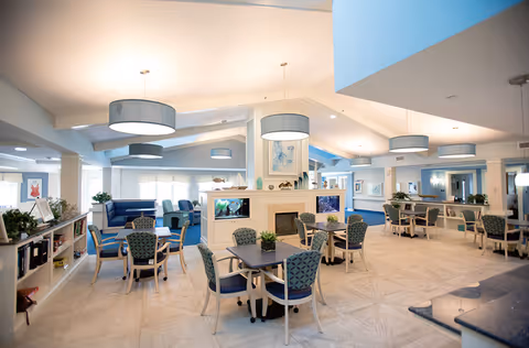 A spacious, well-lit common area in a senior living facility featuring multiple seating arrangements with tables and chairs. The room has a central fireplace with built-in aquariums on either side, modern pendant lights hanging from the ceiling, and large windows allowing natural light to fill the space. The decor includes plants, artwork, and a mix of blue and neutral tones.
