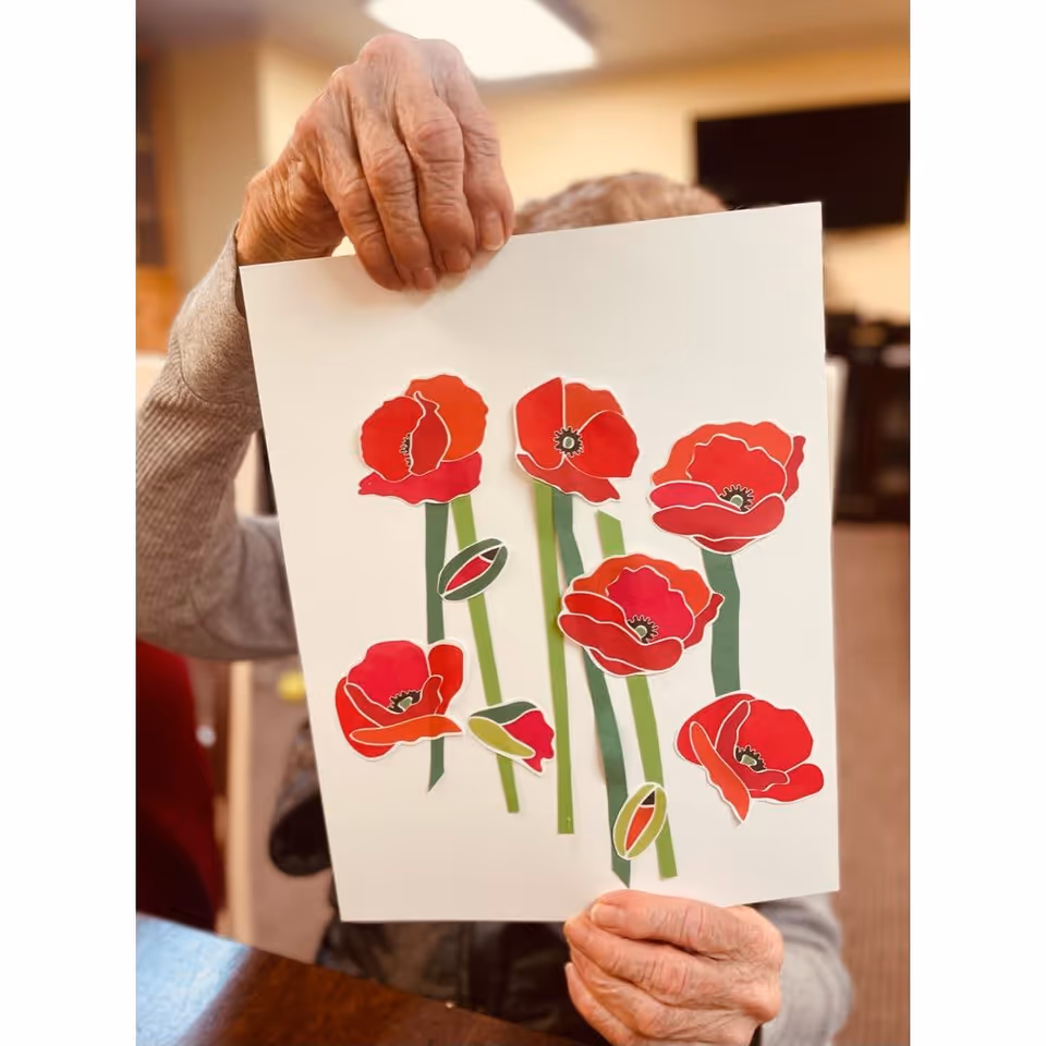 An elderly person holding up a piece of white paper with a colorful paper craft of red poppy flowers with green stems. The person's face is mostly obscured by the paper, and the background shows an indoor setting with warm lighting.