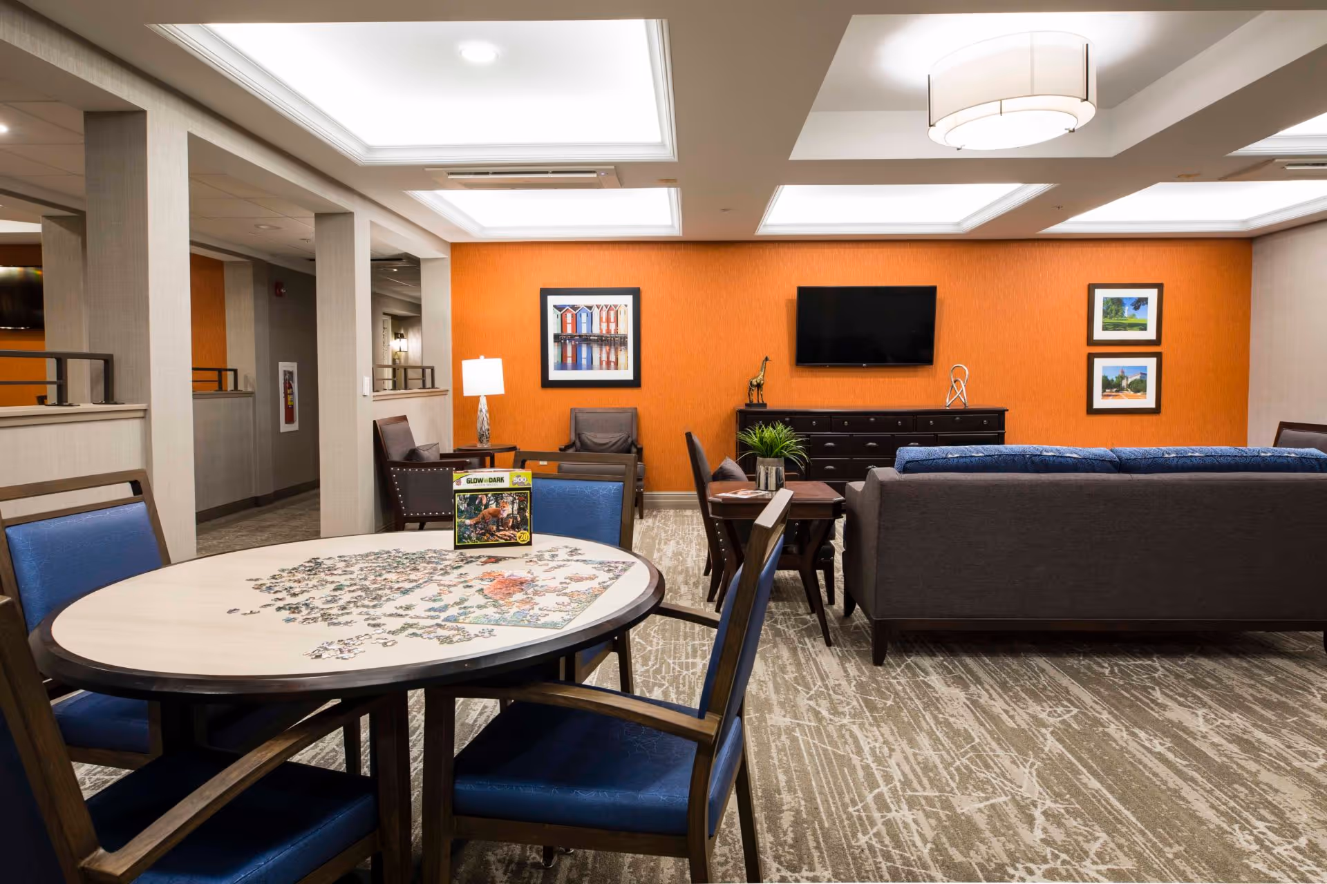 A communal living room/lounge with a round table and puzzle in the foreground, seating areas, and a wall-mounted TV on an orange accent wall.