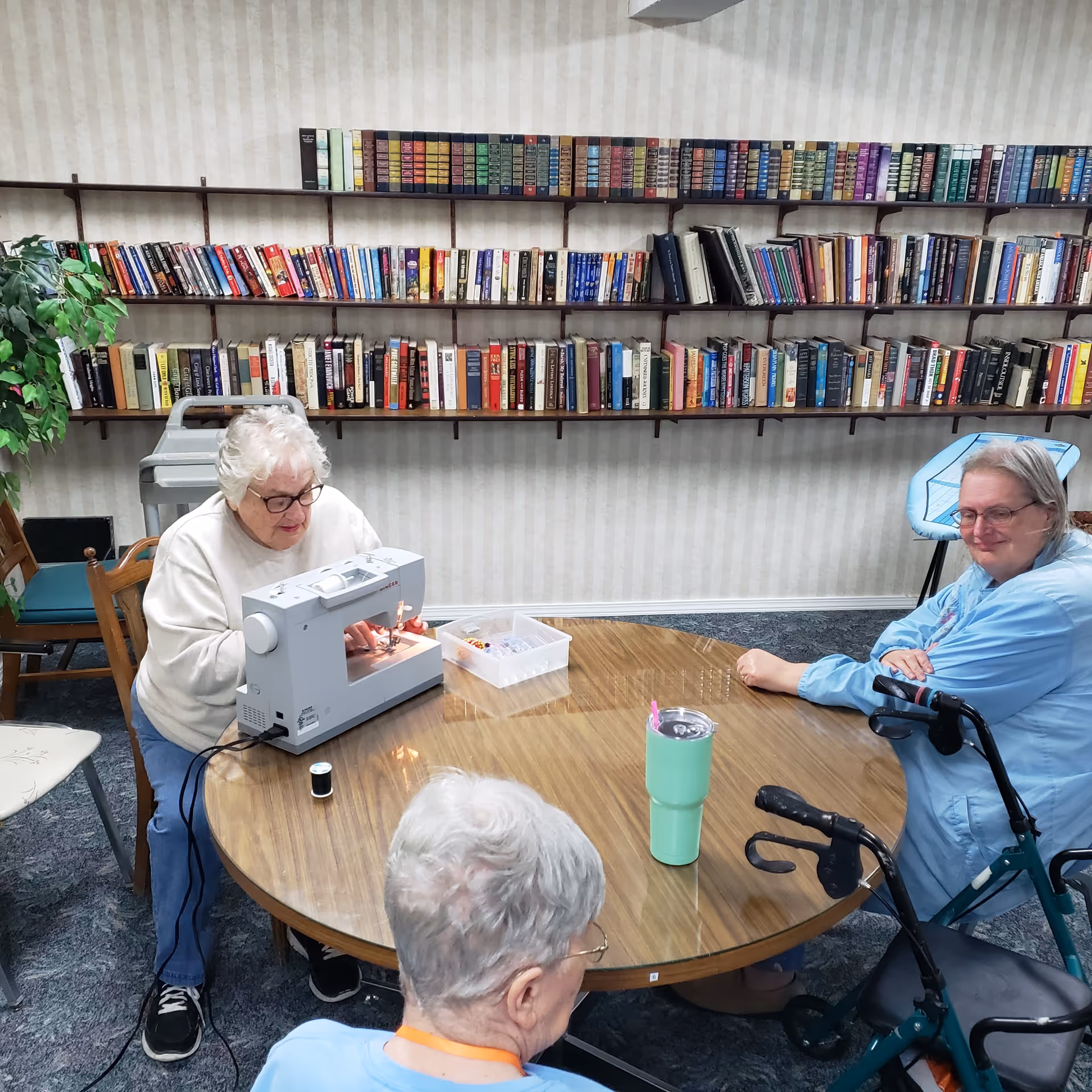 Three elderly women sitting around a round wooden table in a room with bookshelves filled with books on the wall behind them. One woman is using a sewing machine, another woman is sitting with a walker nearby, and the third woman is seated with her back to the camera. There is a green tumbler on the table and a plant in the corner of the room.