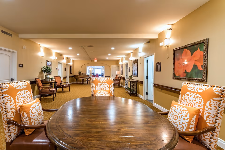 A well-lit communal dining area and hallway with a wooden table and orange patterned upholstered chairs in a senior living facility.
