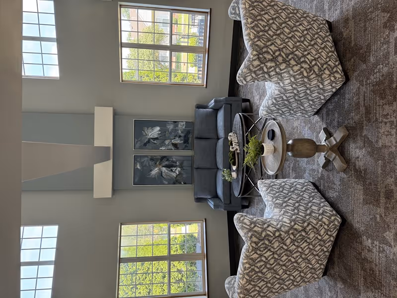 A bright and spacious living room area with large windows letting in natural light. The room features a gray sofa, two patterned armchairs, a round coffee table with decorative plants, and two floral paintings on the wall. The carpet has a textured pattern, and the walls are painted in a light neutral color.