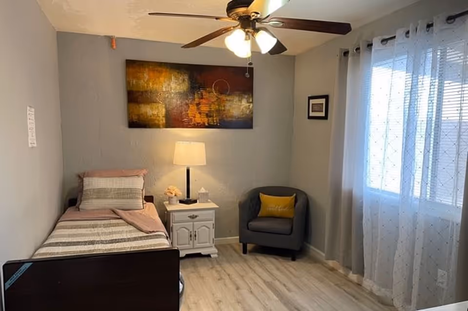 A small, cozy bedroom with a single bed on the left side, covered with a striped blanket and a pillow. Next to the bed is a white nightstand with a lamp, a tissue box, and a small decorative item. Above the nightstand hangs an abstract painting with warm colors. To the right, there is a gray armchair with a yellow pillow. The room has light-colored wooden flooring, light gray walls, a ceiling fan with lights, and a window with sheer white curtains allowing natural light to enter.