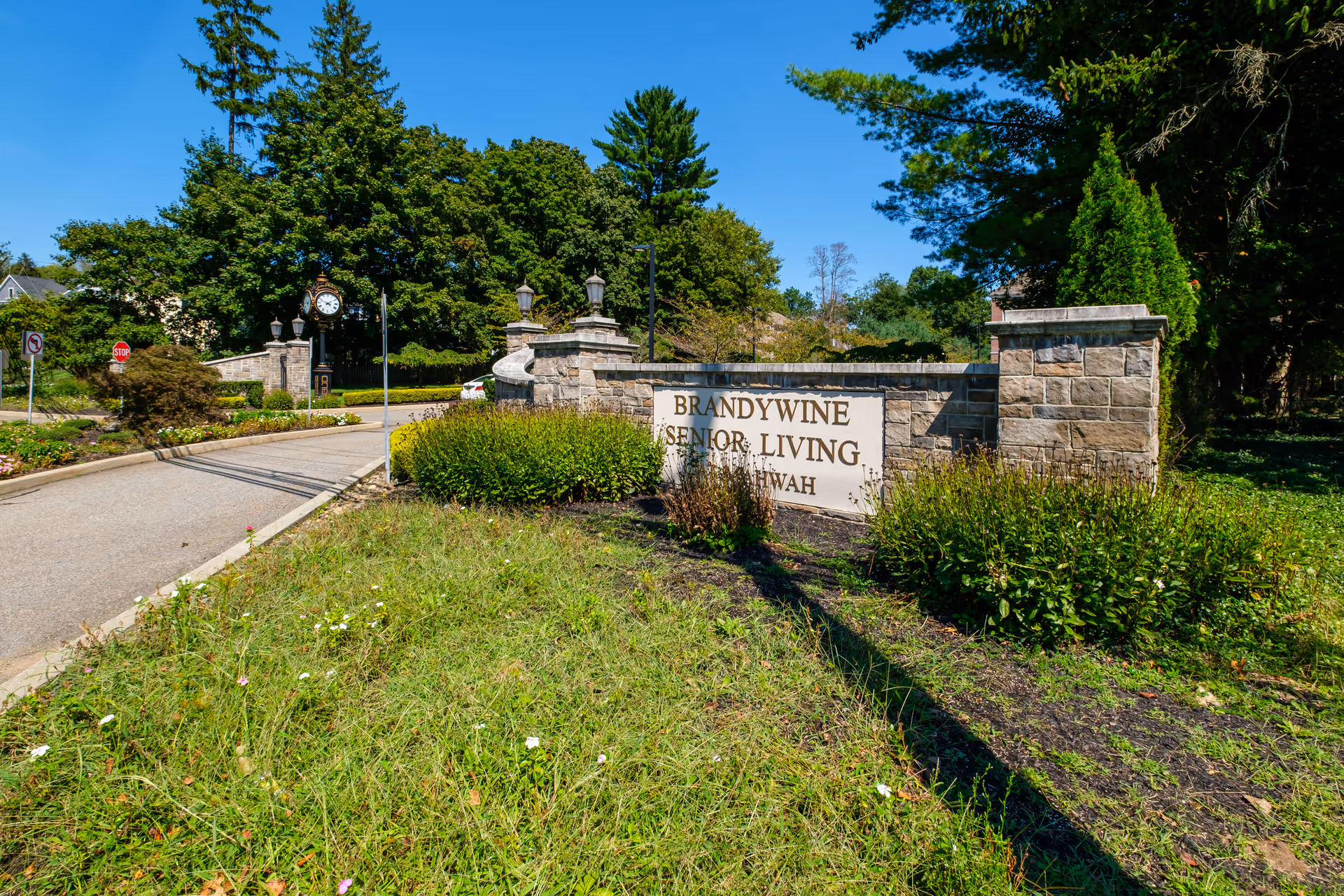 Entrance sign for Brandywine Senior Living Mahwah made of stone with bushes and greenery around it, a paved road leading into the facility, and trees in the background under a clear blue sky.