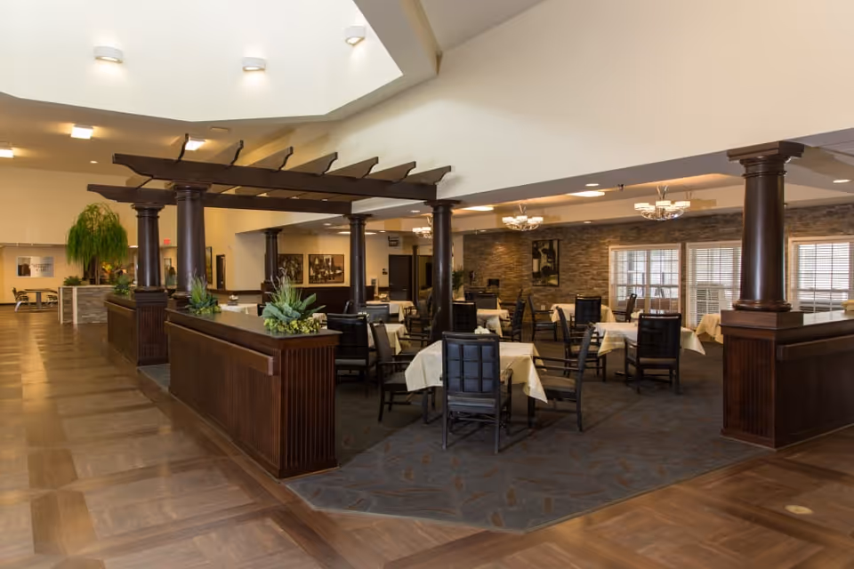 Interior view of a senior living facility dining room with multiple tables covered with white tablecloths and surrounded by dark wooden chairs. The area is decorated with wooden columns and a pergola-style overhead structure. The floor has a combination of wood and carpet, and the walls feature stone accents and windows with blinds. Ceiling lights provide warm illumination.