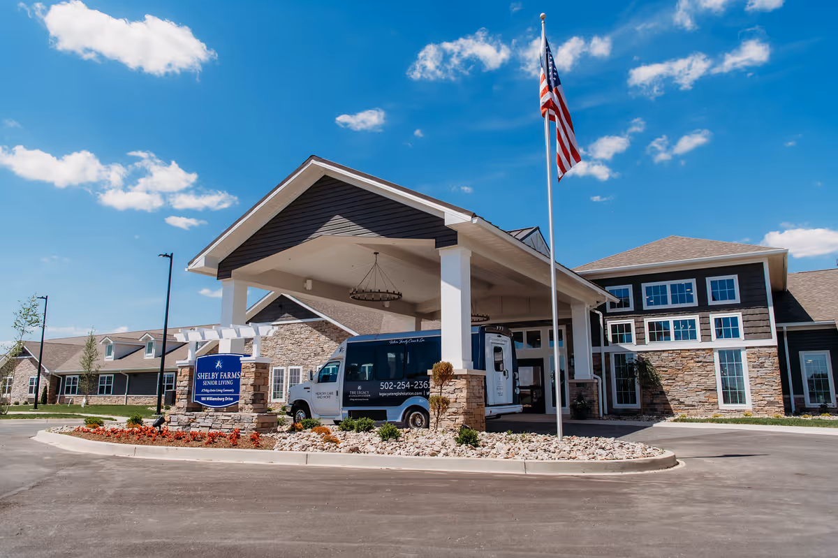 Front entrance of Shelby Farms Senior Living with a covered porte-cochère, shuttle van, flagpole, and property sign.