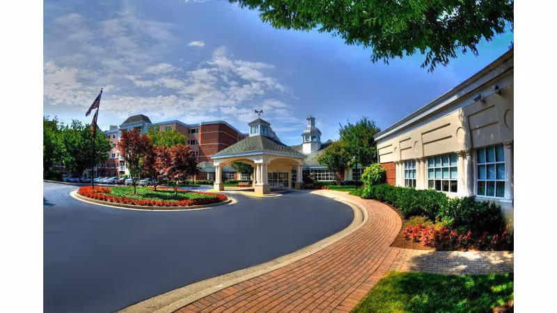 Exterior view of a senior living facility named Blakehurst featuring a circular driveway with a covered entrance, landscaped flower beds, trees, and a clear blue sky.