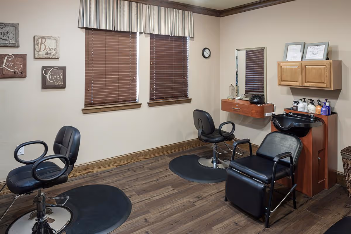 Small salon room with three styling chairs, a shampoo sink, wall cabinets, and two windows with blinds.