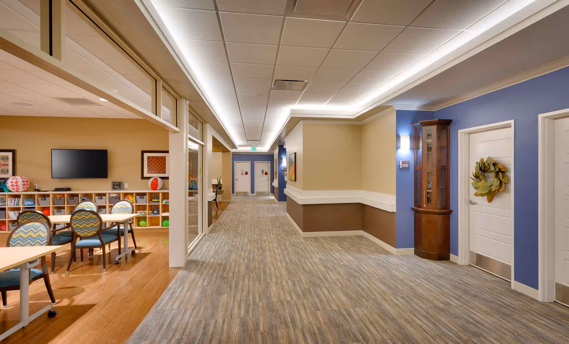 Wide, well-lit interior hallway of a senior living facility with an activity/seating area to the left and doors along the right.