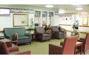 A well-lit common area in a senior living facility with multiple upholstered chairs and sofas arranged around the room. The walls are decorated with framed artwork and floral wallpaper borders. There are plants and floral arrangements placed on tables and shelves, creating a welcoming atmosphere.
