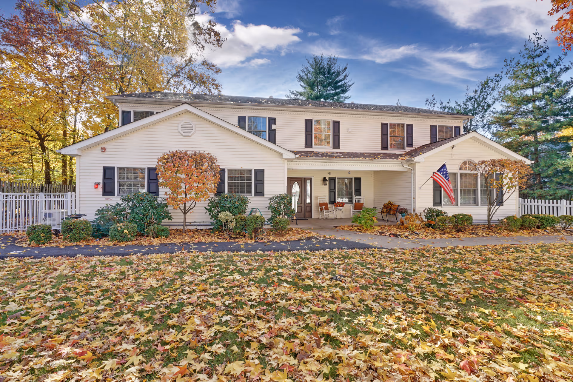 A two-story white building with black shutters surrounded by autumn trees and fallen leaves on the grass. There is a small porch with chairs and an American flag mounted near a window. The sky is partly cloudy with blue patches.