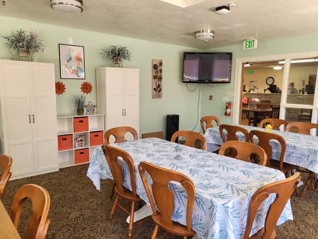 A dining room in an assisted living facility with tables covered in blue and white patterned tablecloths, surrounded by wooden chairs. The room has light green walls decorated with plants, framed artwork, and wall hangings. There are white storage cabinets and a wall-mounted TV. A doorway leads to another room with more tables and chairs visible.