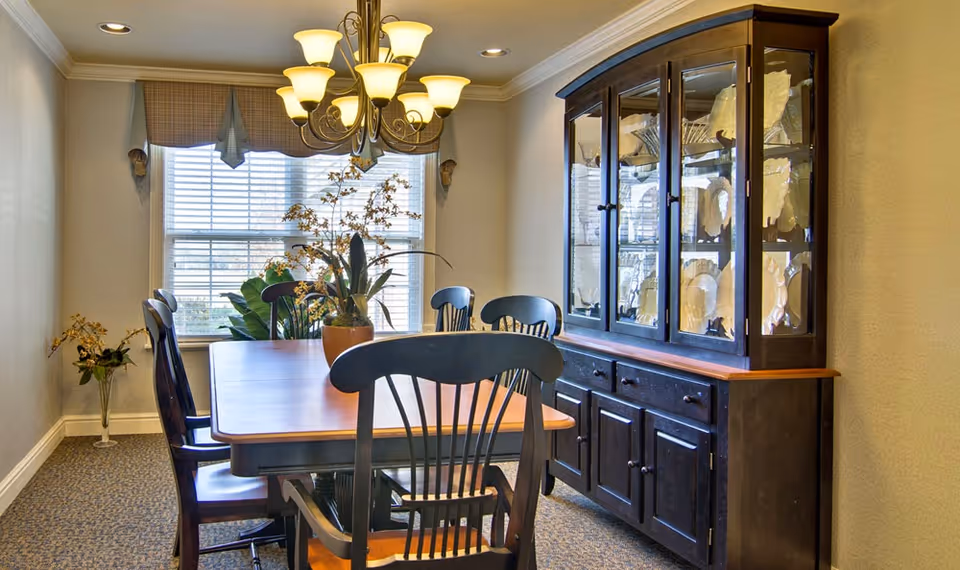 A dining room with a wooden dining table surrounded by six black wooden chairs. A chandelier with multiple lights hangs above the table. There is a large window with blinds and a valance, and a dark wooden china cabinet filled with white dishes against the right wall. A potted plant with flowers is placed on the table, and another plant is visible near the window.