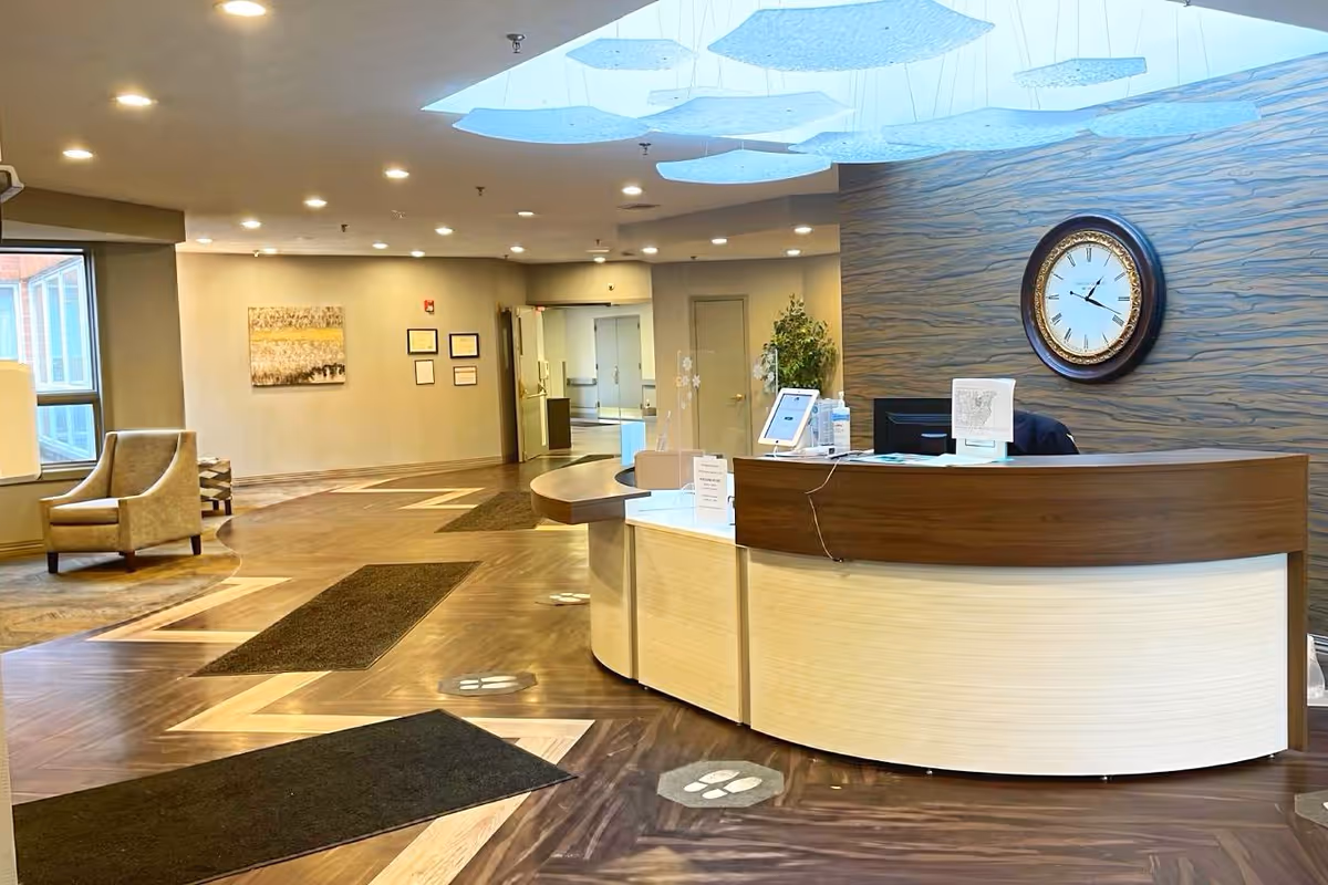 Reception area of a senior living facility with a curved wooden front desk, a large wall clock, modern ceiling lights, and seating area with armchairs near a window.