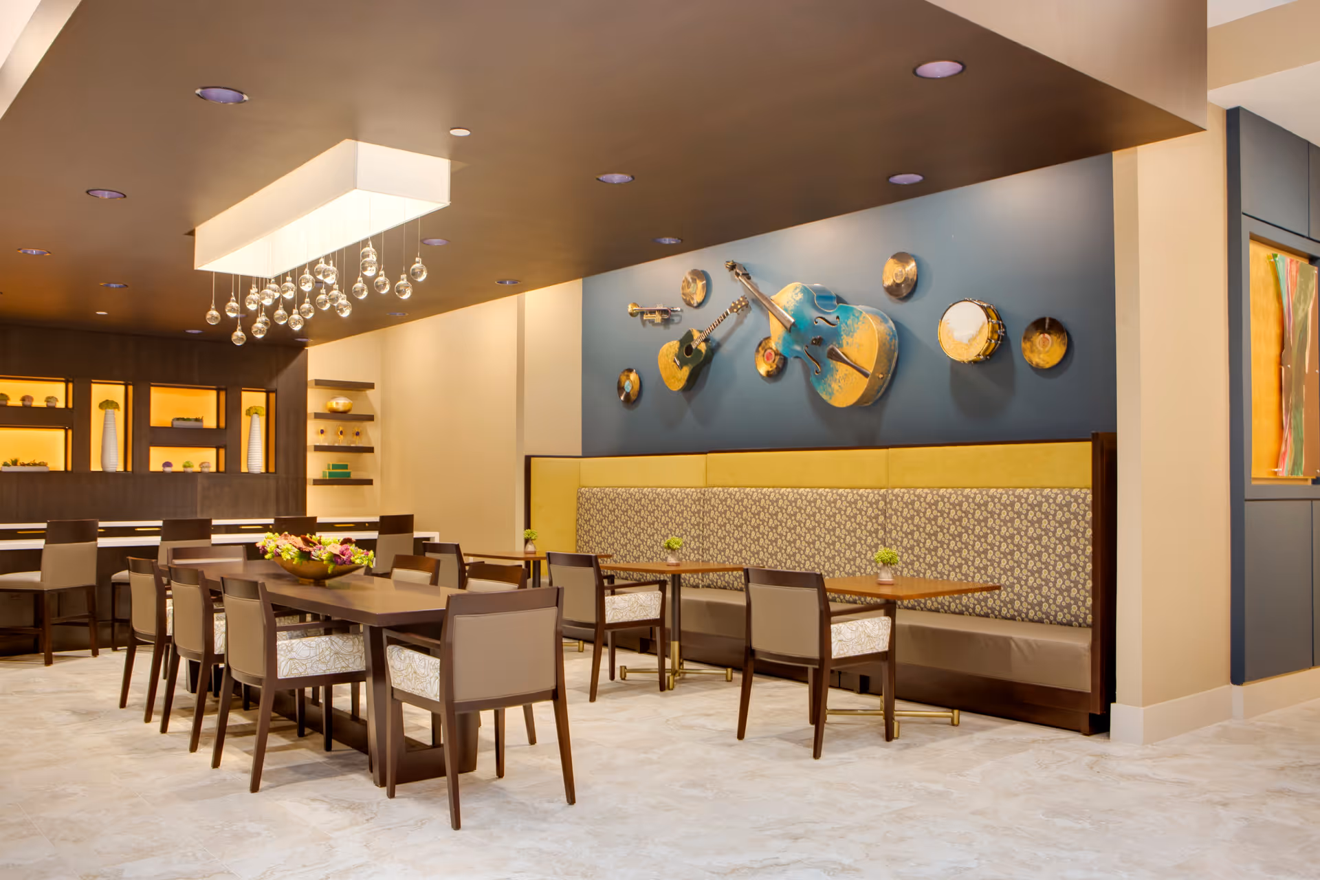 A modern dining room with a long wooden table surrounded by chairs, a built-in bench with patterned upholstery along the wall, and decorative musical instruments mounted on a blue accent wall. The room features a contemporary chandelier with hanging glass orbs and built-in shelving with decorative items.