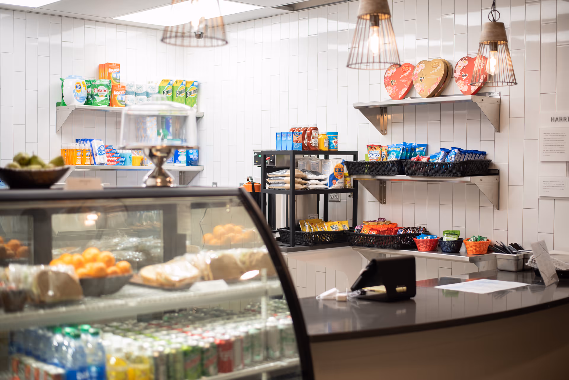 A snack shop counter with a glass refrigerated display case, shelves stocked with packaged snacks and drinks, and hanging pendant lights.