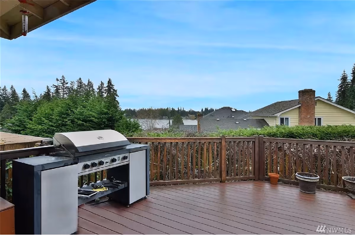 Wooden deck with a stainless steel gas grill, a few potted plants, and a view of neighboring houses, trees, and a distant lake.