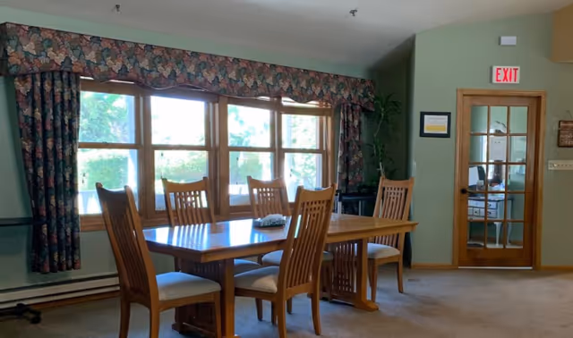 Wooden dining table with six chairs in a sunlit room featuring large windows with floral curtains and a glass door with an EXIT sign.
