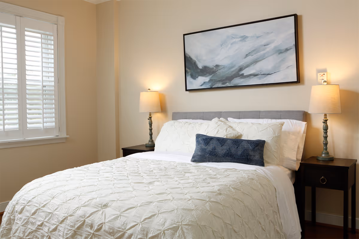 A neatly made bed with white textured bedding and multiple pillows, including a dark blue decorative pillow. The bed has a gray upholstered headboard and is flanked by two dark wooden nightstands, each with a lamp. Above the bed hangs a large abstract painting in shades of gray and white. To the left, there is a window with white plantation shutters.