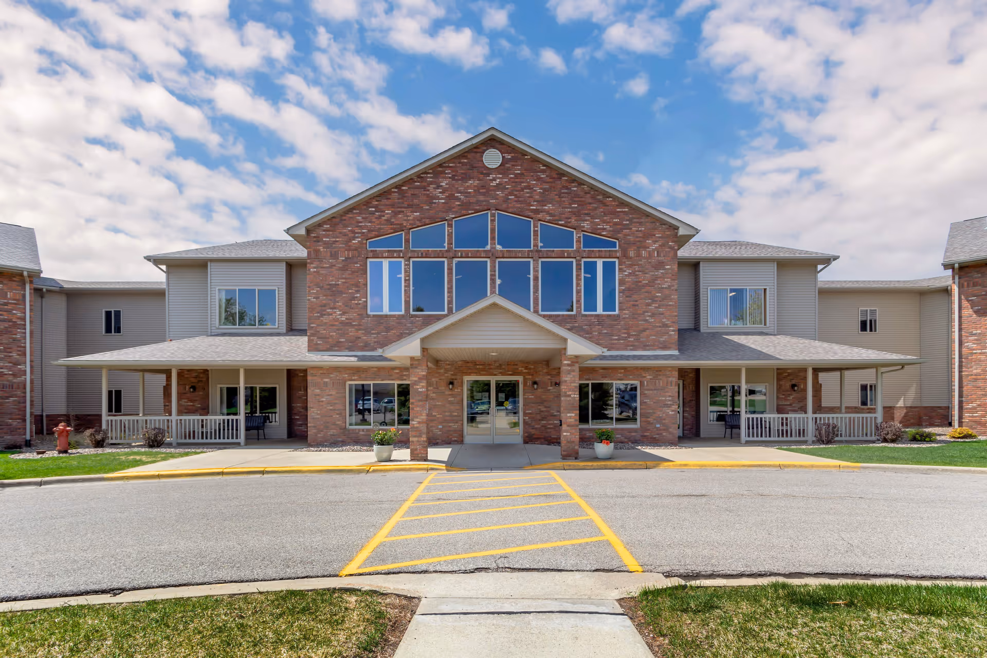 Front exterior view of South Grove Lodge Senior Living building with brick and siding facade, large windows, covered entrance, and a parking area with yellow striped lines in front.